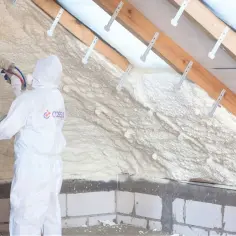 Osoba w kombinezonie wykonuje klejenie styropianu na piankę, izolując poddasze.