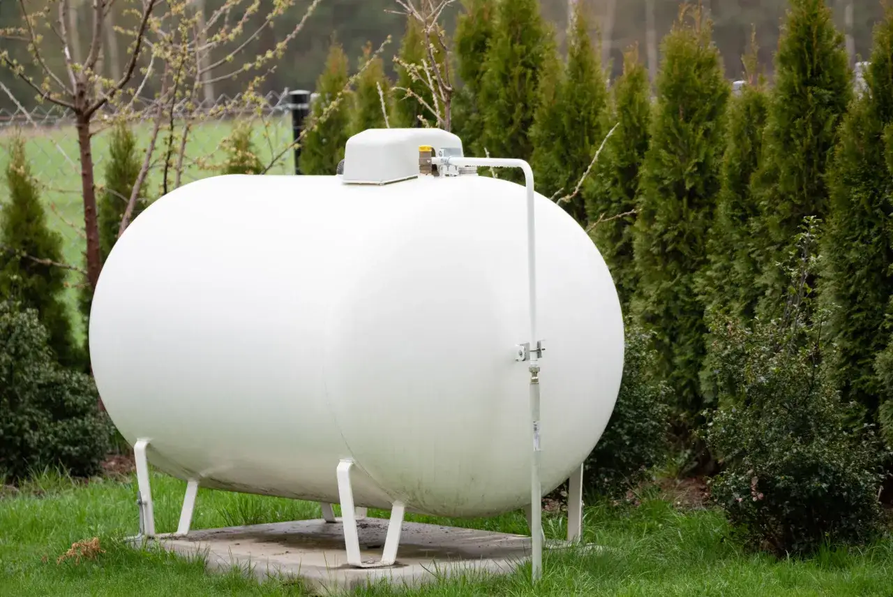 Duży, biały zbiornik na gaz 2700 litrów stoi na trawniku, otoczony zielenią.